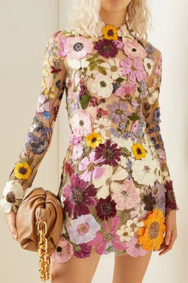 Close-up of a sheer mini dress with colorful floral embroidery and appliqués, held by a woman with a brown clutch.