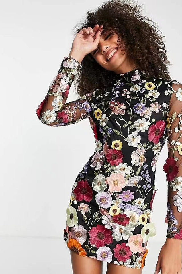 Woman with curly hair smiling, wearing a black sheer mini dress covered in colorful floral embroidery.