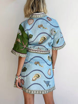 The back of a short-sleeved shirt dress featuring a wavy blue background with various shells and fish printed on it. The dress has a green panel on the left shoulder.