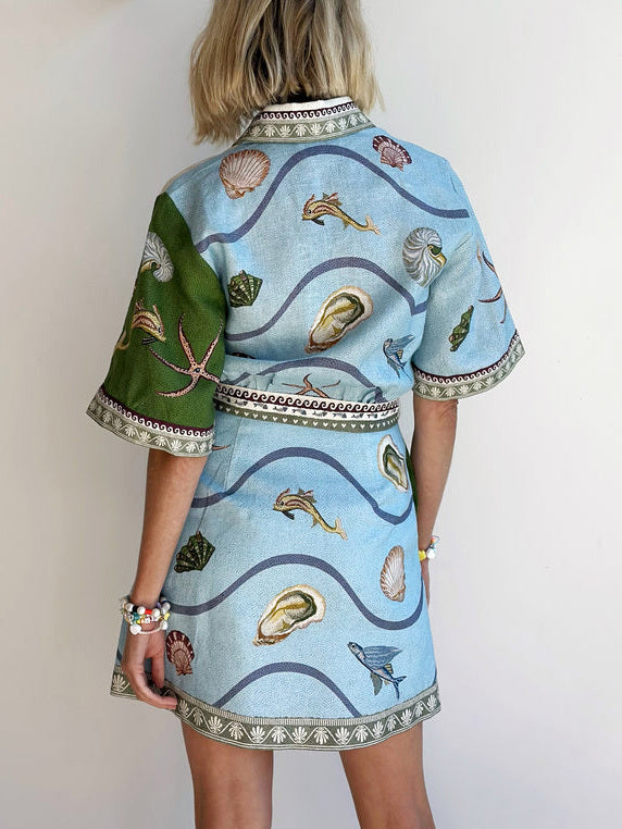 The back of a short-sleeved shirt dress featuring a wavy blue background with various shells and fish printed on it. The dress has a green panel on the left shoulder.