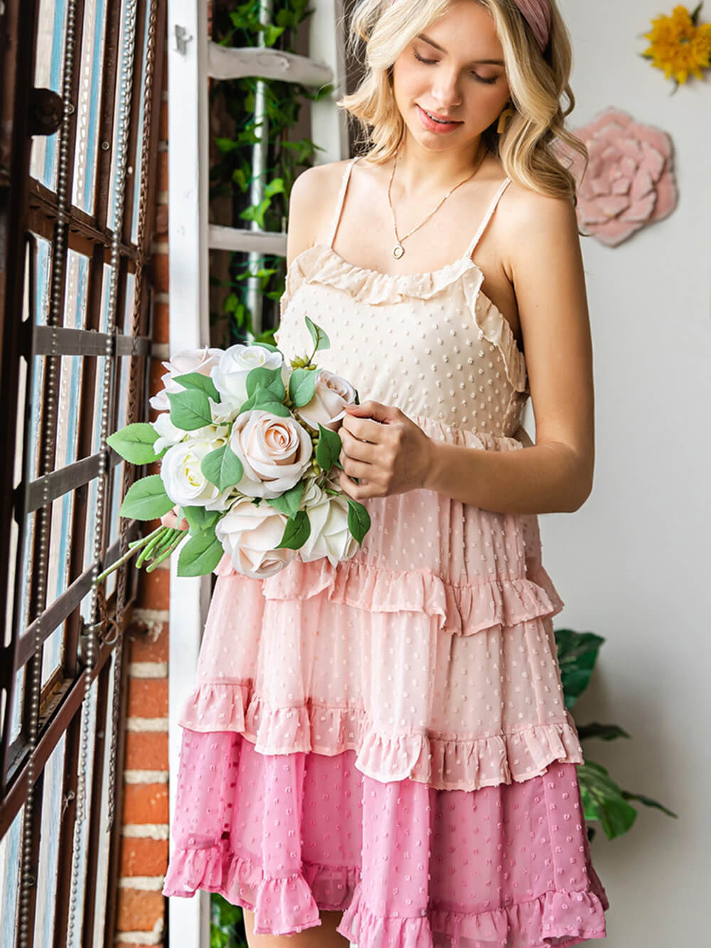 A young woman holding a bouquet of flowers, wearing a pink and beige ruffled dress with a swiss dot pattern, standing near a window.