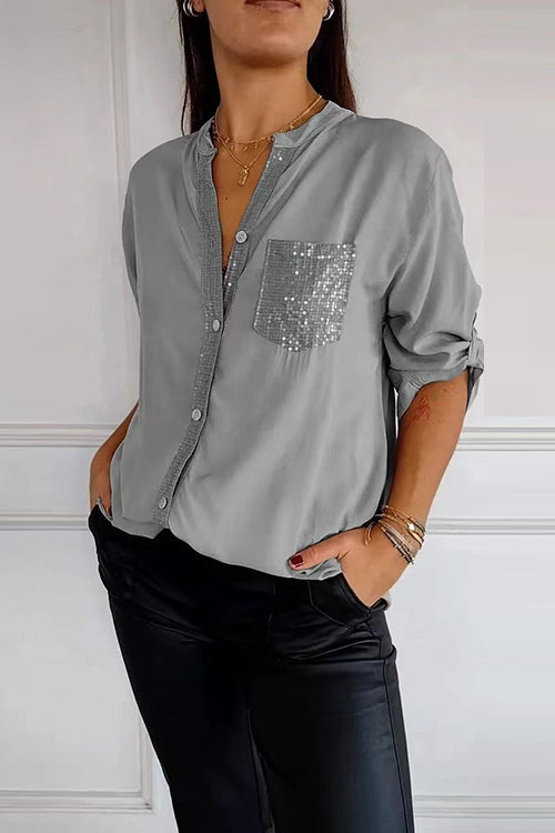 A woman wears a light grey long-sleeved shirt with a sequined pocket and black pants.