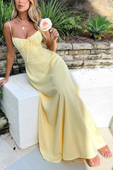 A woman in a yellow maxi dress with spaghetti straps and a tie-front bodice, sitting on a ledge in a garden.