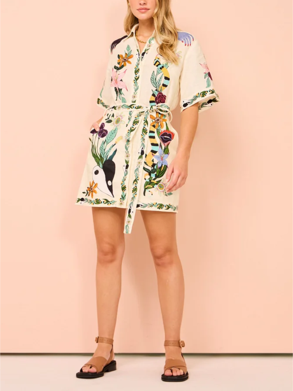 Full-body shot of a woman wearing a cream-colored short-sleeve shirt dress with a colorful floral and animal print, belted at the waist.