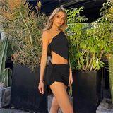 A young woman stands in front of lush greenery wearing a black one-shoulder top and a matching mini skirt.