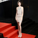 A model stands on a red carpet wearing a short, white sequined halter neck dress with fringe.