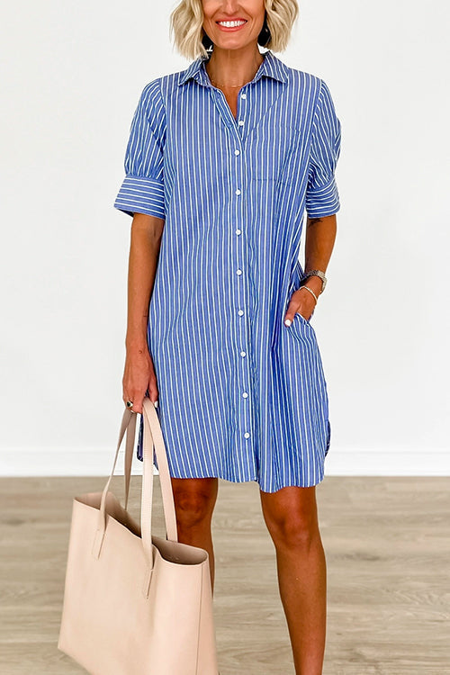 Close-up of a woman wearing a blue and white striped shirt dress with her hands in the pockets and holding a large beige tote bag.