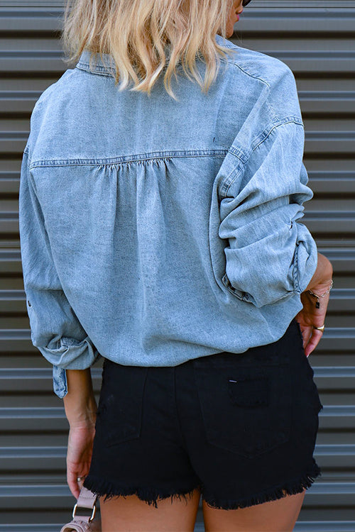Back view of a woman in a light blue cropped denim shirt and black distressed shorts.