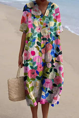 A woman wearing a colorful floral print midi dress with a v-neck and short sleeves, holding a straw tote bag on a beach.