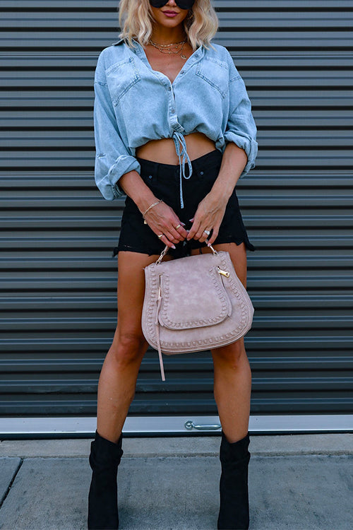 Full body shot of a woman in a light blue cropped denim shirt, black distressed shorts, black boots, and a beige handbag.