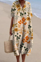A woman wearing a cream-colored midi dress with an orange and green floral print, v-neck, and short sleeves, holding a straw tote bag on a beach.