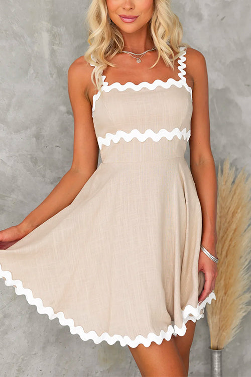 Front view of a woman wearing a beige fit and flare mini dress with white scalloped trim on the straps and hem.