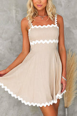 Front view of a woman wearing a beige fit and flare mini dress with white scalloped trim on the straps and hem.