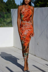 A woman modeling an orange and brown tie-dye print ruched halter neck midi dress with strappy heels, standing outdoors.