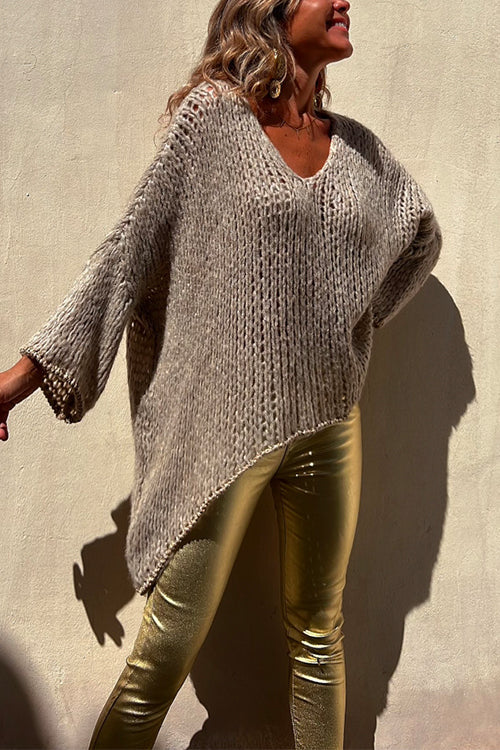 Woman wearing a beige oversized knit sweater with gold metallic pants, standing against a wall.