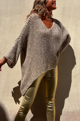 Woman wearing a beige oversized knit sweater with gold metallic pants, standing against a wall.