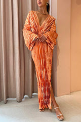 Woman wearing an orange pleated maxi dress with a deep V-neckline and long sleeves, posing in front of a beige curtain.