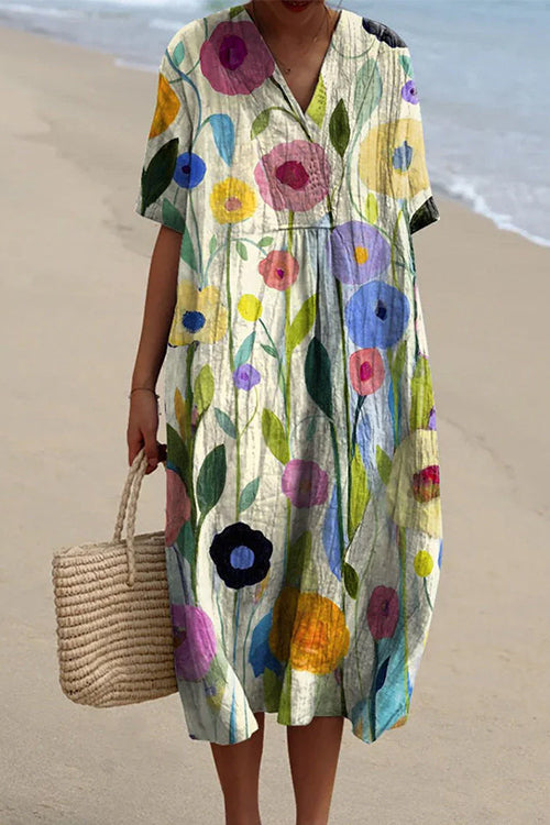 A woman wearing a cream-colored midi dress with a vibrant floral print featuring pink, blue, and yellow flowers, v-neck, and short sleeves, holding a straw tote bag on a beach.