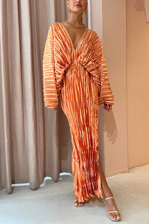 Woman wearing an orange pleated maxi dress with a deep V-neckline and long sleeves, standing in front of a beige curtain.