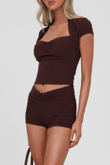 woman wearing a brown ruched crop top with a matching bolero jacket and shorts, on a white background