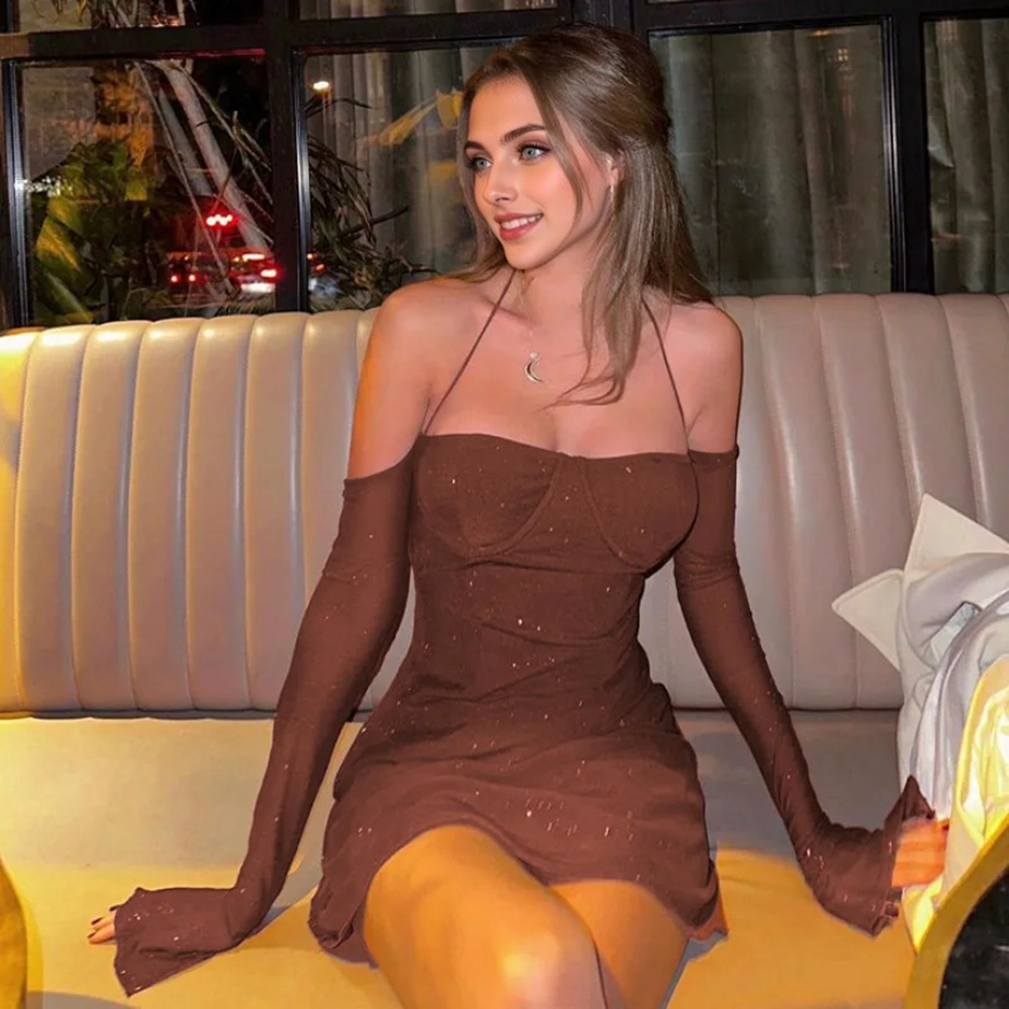 A young woman with blonde hair pulled back and blue eyes smiles while sitting on a plush couch in a short, brown, sparkly dress with off-the-shoulder sheer sleeves and a halter neckline. She is wearing a moon necklace.