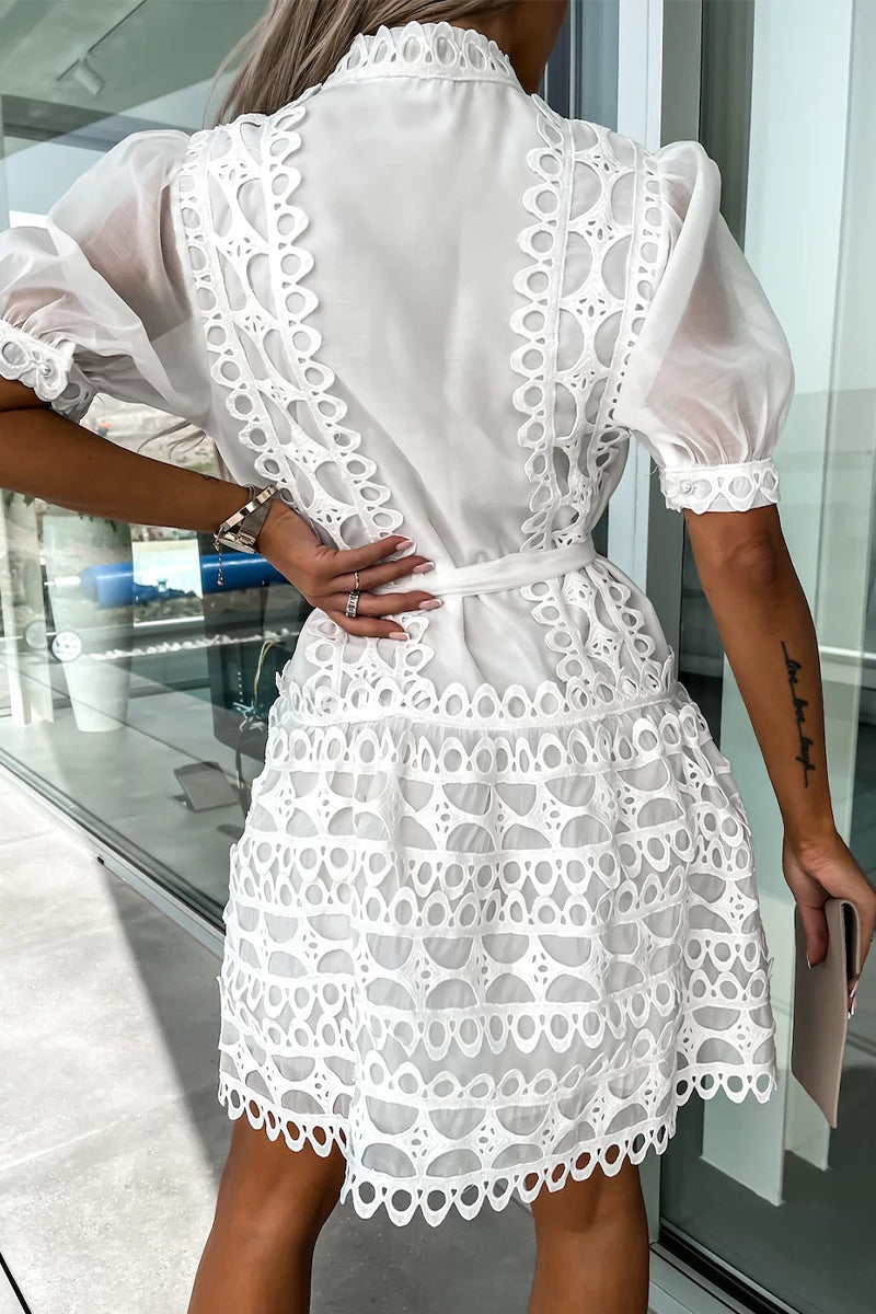 Woman wearing a white lace dress with puff sleeves and a belted waist, holding a beige clutch.