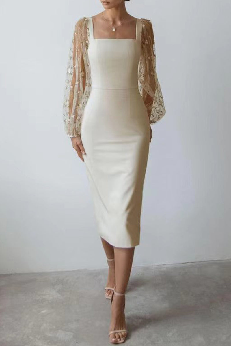 A woman stands against a white wall wearing a cream-colored sheath dress with sheer, detailed sleeves.