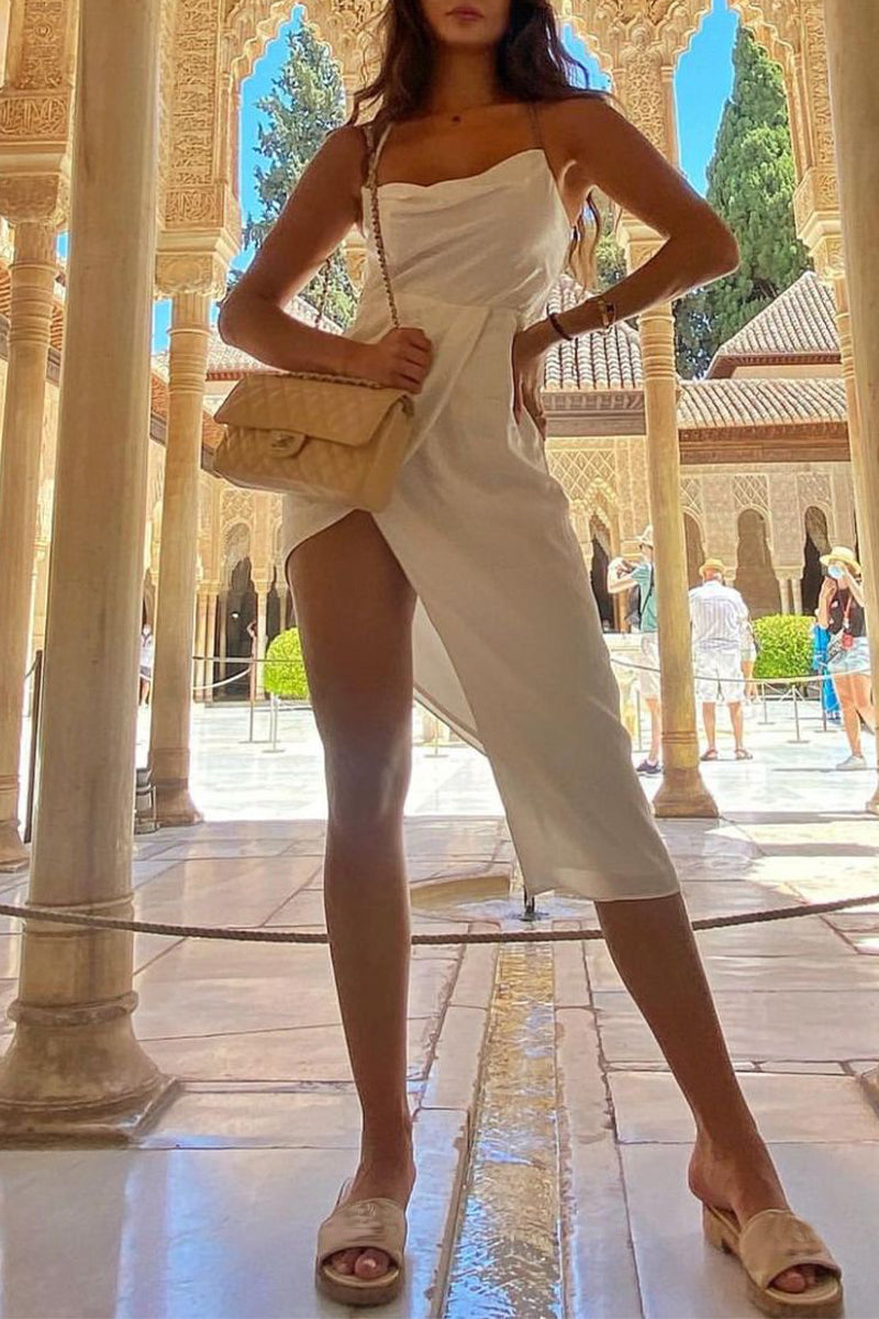 A woman poses in a white cowl neck dress with a high slit, accessorized with a gold belt and a beige handbag, standing in an ornate courtyard.