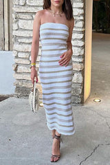 A woman in a white and tan striped strapless knit dress and black strappy heels holds a white net bag.