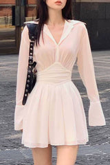 A woman wearing a light pink sheer dress with a cinched waist and flared sleeves, standing outdoors.