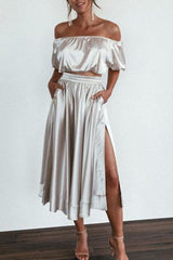 A woman models a silver satin off-the-shoulder crop top and a matching high-waisted midi skirt with a side slit.