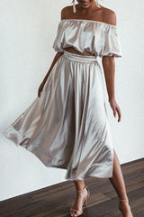 A woman in a silver satin outfit with an off-the-shoulder crop top and a high-waisted midi skirt with a side slit.