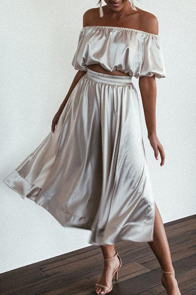 A woman in a silver satin outfit with an off-the-shoulder crop top and a high-waisted midi skirt with a side slit.