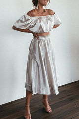 A woman poses with her hands on her hips, wearing a silver satin off-the-shoulder crop top and a matching midi skirt with a side slit.