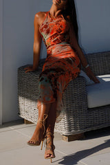 A woman sitting on an outdoor couch, modeling an orange and brown tie-dye print ruched halter neck midi dress with strappy heels.