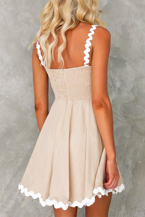 Back view of a woman wearing a beige fit and flare mini dress with white scalloped trim on the straps and hem.
