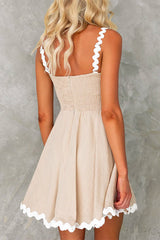 Back view of a woman wearing a beige fit and flare mini dress with white scalloped trim on the straps and hem.