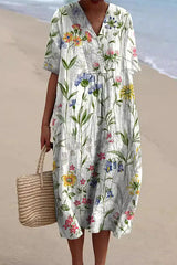 A woman wearing a white midi dress with a delicate floral print in shades of blue, pink, and yellow, v-neck, and short sleeves, holding a straw tote bag on a beach.