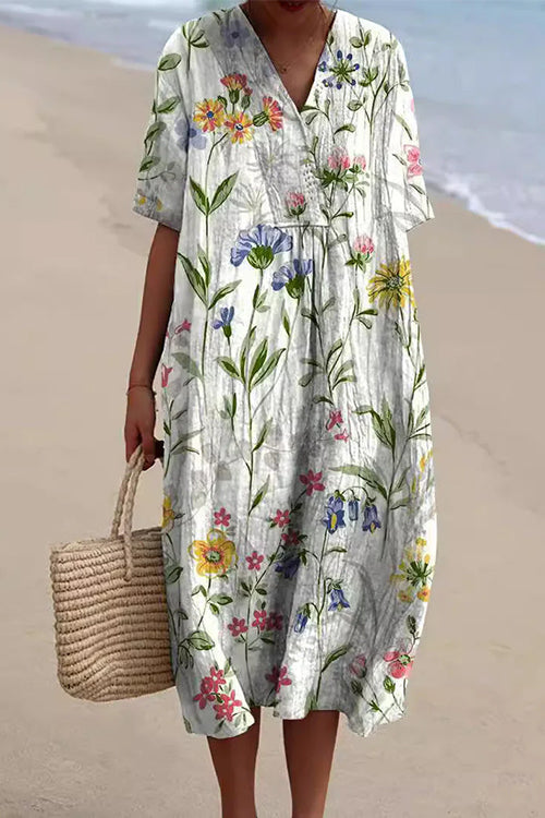A woman wearing a white midi dress with a delicate floral print in shades of blue, pink, and yellow, v-neck, and short sleeves, holding a straw tote bag on a beach.