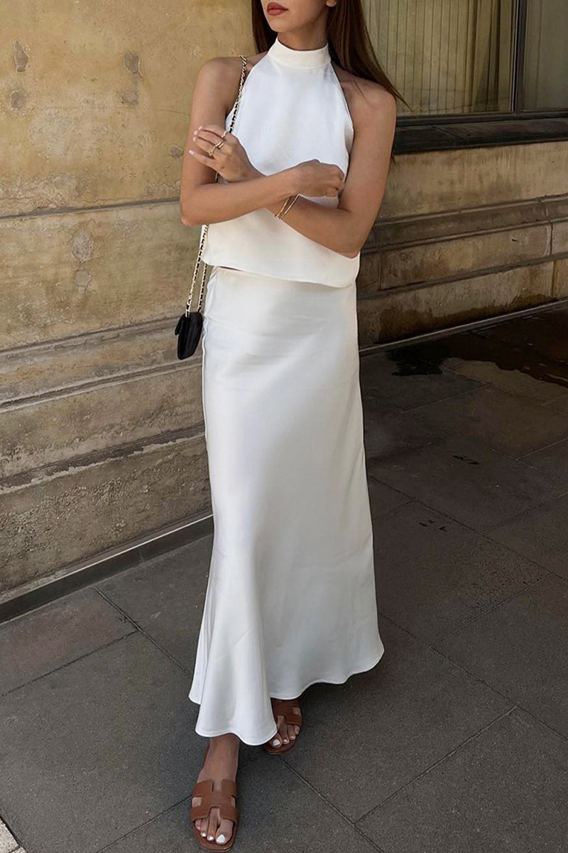 A woman in a white halter top and a flowing white satin maxi skirt with arms crossed.