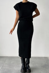 A woman wearing a black ribbed knit sleeveless top and a black ribbed knit midi skirt with black boots.