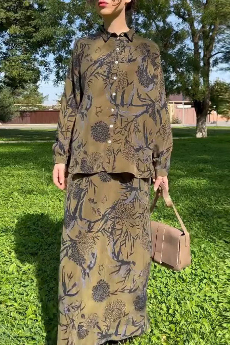 Woman wearing a long-sleeved olive green button-down shirt with a black and gray floral print, paired with a matching long skirt. She is holding a beige handbag and standing in a grassy park.
