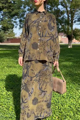 Woman wearing a long-sleeved olive green button-down shirt with a black and gray floral print, paired with a matching long skirt. She is holding a beige handbag and standing in a grassy park.