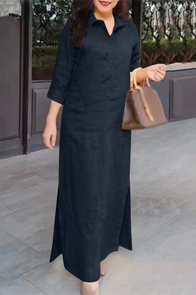 A woman stands wearing a long black dress with a collared neckline and three-quarter sleeves.