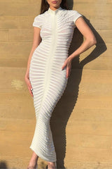 Woman in a white ribbed bodycon maxi dress with a high neckline and short sleeves, posing against a wooden wall.