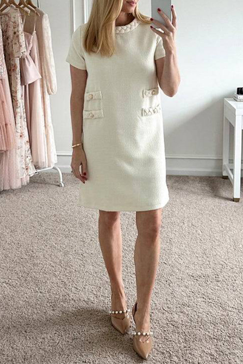 Woman wearing a cream textured A-line shift dress with short sleeves, a ruffled neckline, and four decorative pockets with button and braided trim. She is also wearing nude pointed-toe heels with pearl embellishments.