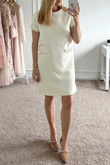 Woman wearing a cream textured A-line shift dress with short sleeves, a ruffled neckline, and four decorative pockets with button and braided trim. She is also wearing nude pointed-toe heels with pearl embellishments.