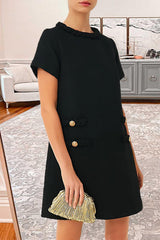 Woman wearing a black textured A-line shift dress with short sleeves, a ruffled neckline, and four decorative pockets with button and braided trim. She is holding a gold clutch.