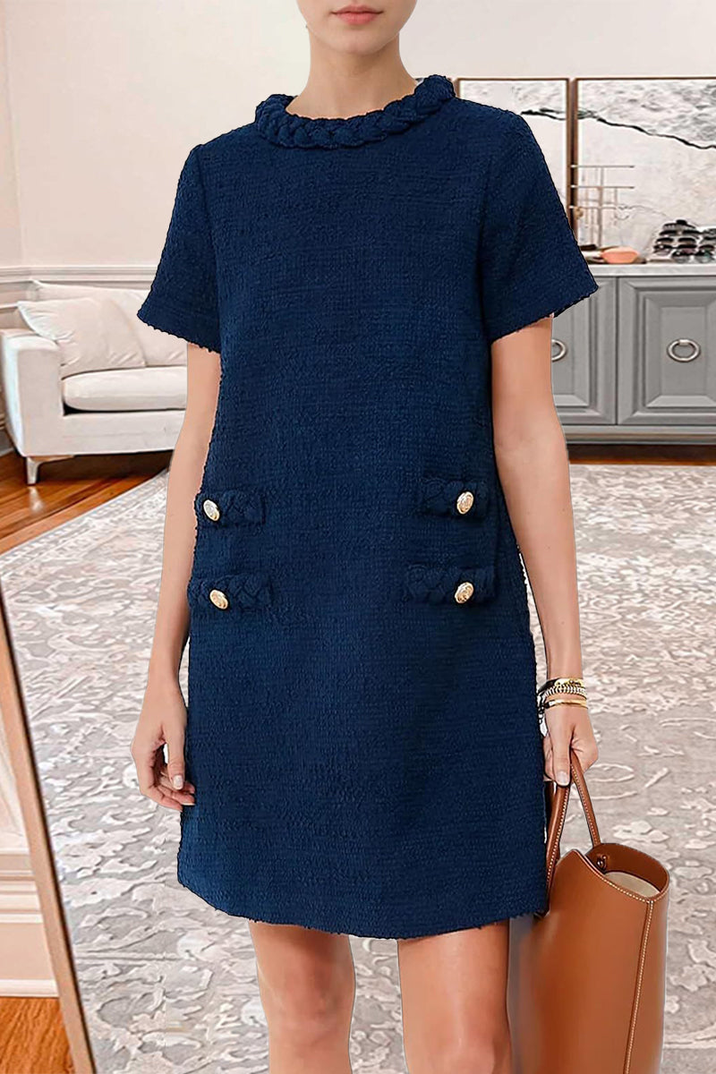 Woman wearing a navy blue textured A-line shift dress with short sleeves, a ruffled neckline, and four decorative pockets with button and braided trim. She is also wearing a brown tote bag and layered bracelets.