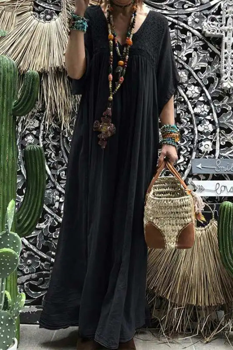 A woman wearing a long, flowing black maxi dress with short sleeves and a v-neckline. She is accessorized with a beaded necklace and bracelets and is carrying a woven bag. The background features cacti and decorative wall art.